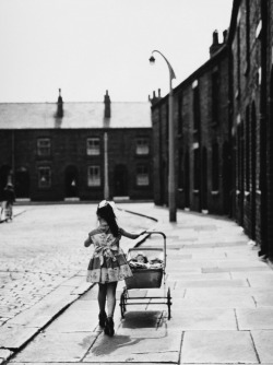 m3zzaluna:  a little girl in a pretty dress wearing high heels which are far too large for her feet, pushes her toy pram along a street in wigan, 1961 © shirley baker » check out more of baker’s photography here
