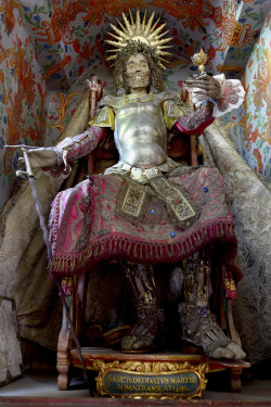 St. Deodatus (Rheinau, Swizterland) “One of two seated skeletons which arrived late in the 17th century in the town’s monastic church. He shows a rare style of facial decorations, with a wax mask molded over the upper half of the skull, and a cloth