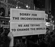 [Person holding up a sign: “Sorry for the inconvenience, we are trying to change the world.”]